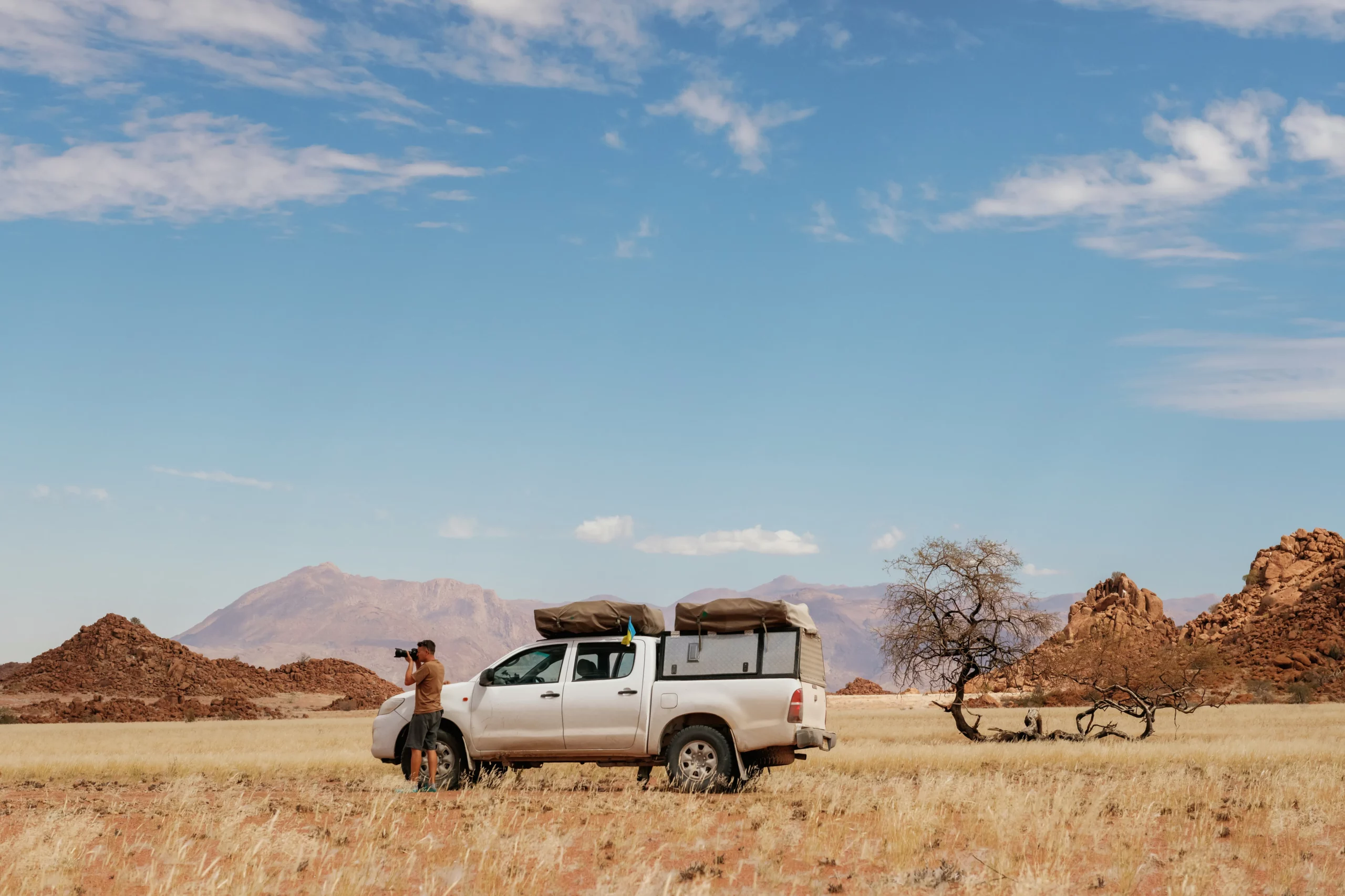 Une personne se tient à côté d'une camionnette blanche et prend des photos dans un paysage désertique sec et herbeux en Namibie. À proximité, un arbre solitaire et des montagnes lointaines sous un ciel bleu créent une atmosphère sereine.